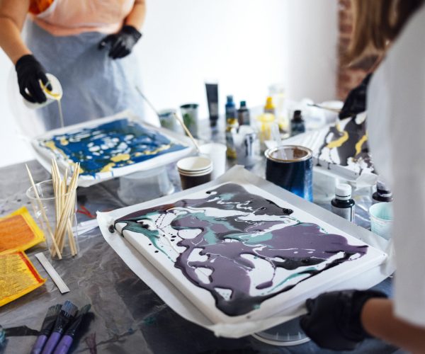 Painting tutorial. Two white women in the gloves paint with liquid acrylic in an art workshop. Acrylic Fluid Pouring. Dirty glass technique. Paintings, paints and brushes on the table. Fluid Art.